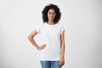 Wall Mural - Stylish young dark-skinned female with curly hair looking at camera and smiling cutely, posing with hand on her waist, isolated on white background with copy space for your promotional content