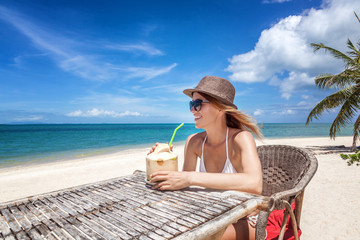 Wall Mural - Happy blonde woman in hat and sunglasses on the tropical beach sitting in cafe with coconut