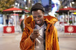 Happy young African American man dressed stylishly in winter coat and hat having evening walk alone on streets of foreign city, messaging friends on electronic gadget. People and modern technology