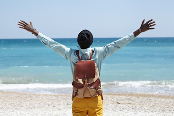 Read view of happy carefree African American male standing on beach in front of azure sea with his back to camera, spreading arms, feeling freedom and connection to amazing nature around him