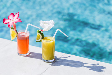 Two glasses of fresh cold fruit juice on poolside. With straw, slice of lime and tropical flower