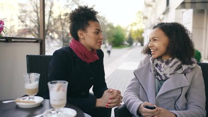 Wall Mural - Two attractive mixed race women talking and drinking coffee in street cafe. Friends have fun after visiting mall sale