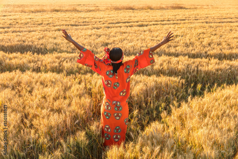 Obraz na płótnie African woman in traditional clothes arms raised in field of crops at sunset or sunrise w sypialni