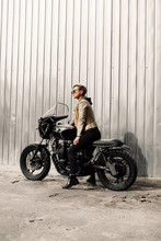 Woman Sitting On Her Motorcycle. Motorcycle Near A Grey Wall. Girl With Short Hair. Girl In Black Glasses And A Leather Jacket. Bike