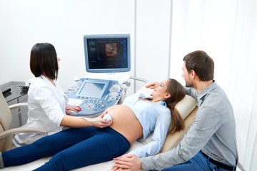 Wall Mural - Happy loving pregnant couple during sonogram research at the fertility clinic. Female doctor performing ultrasound scanning for her pregnant patient sonogram medicine modern technology equipment.