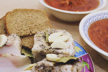 Salted lard with pepper and bay leaf and black bread lies on a plate, next to it there are two plates with red borsch.. Close-up, selective focus.