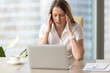 © fizkes - Tired businesswoman suffering of headache. Female entrepreneur sitting at the table in front of laptop and massaging temples. Stressed girl feeling sharp pain in head while working at office
