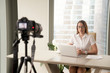 © fizkes - Confident businesswoman sitting at desk in front of laptop and looking at camera on tripod that records video. Female blogger makes footage for social networks. Woman recording videolog at workplace.
