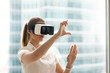 © fizkes - Beautiful girl using virtual reality glasses near bright window with skyscraper view outside. Business woman wearing VR goggles and interacts with cyberspace using swipe and stretching gestures