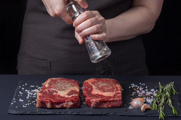 A woman cooks with peppers two fresh raw ribeye steaks from marbled beef on a dark background. Nearby is a mixture of peppers, sea salt, garlic and rosemary