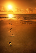 Single Footsteps On Beach Walking Towards Sun Of Coastal Horizon.