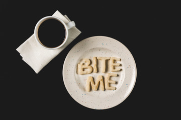 Wall Mural - top view of lettering bite me made from cookie dough on plate with coffee cup isolated on black