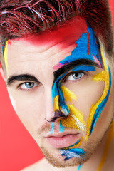 Wall Mural - portrait of young man with colored face paint on red background. Professional Makeup Fashion. fantasy art makeup