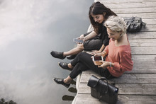 High Angle View Of Happy Businesswomen Using Smart Phone While Sitting On Pier By Lake