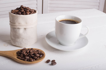 Cup of coffee, coffee beans in the bag, wooden spoon on a wooden white background table. Side view. Place for text or inscription.