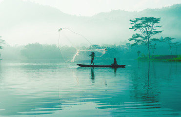 Fisherman casting out his fishing net in the river by throwing it high up into the air early in the blue colored morning to catch fish with his little fishing boat.