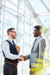 © pressmaster - Portrait of smiling businessman greeting African-American partner by handshake standing in modern glass hall of office building