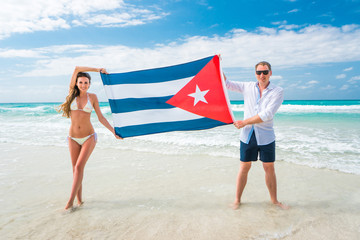 Poster - Beautiful young blond woman in white bikini swimsuit and man are holding the cuban flag in their arms on beach of caribbean sea