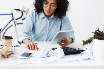 Wall Mural - Serious and concentrated young dark-skinned woman entrepreneur wearing spectacles using digital tablet and calculator while planning company's budget, working through financial papers in her office