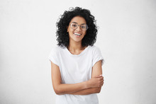 Charming Young Dark-skinned Woman With Curly Hairstyle Having Shy Smile Posing In Studio In Closed Posture, Keeping Arms Folded, Feeling Constrained And A Bit Nervous. Human Emotions And Feelings