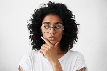 Wall Mural - Education, people and life goals. Headshot of attractive mixed race schoolgirl wearing stylish round eyeglasses having doubtful and indecisive look, holding her chin as she has to choose college