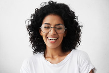 Pretty Girl With Dark Clean Skin And Beautiful Toothy Smile Laughing Out Loud At Funny Joke While Having Fun Indoors With Friends, Closing Eyes In Joy, Looking Carefree And Relaxed. Horizontal
