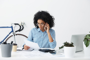 Serious concentrated young female entrepreneur talking on mobile phone with bank concerning sheet of paper in her hand, having frustrated look while working through finances in modern office