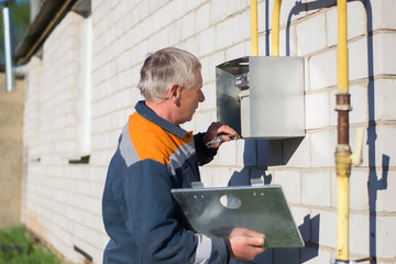 A gray-haired master specialist man repairs a gas meter hanging on a brick wall. Maintenance of gas equipment