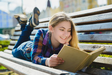 Little Pretty Girl Reading A Book And Sitting On Bench Outdoor