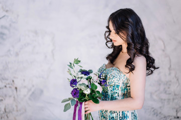 beautiful sexy Oriental girl with makeup black curls in a bright green sequined dress with purple and pink bouquet on his hand looking down profile on white gray background, horizontal copyspace