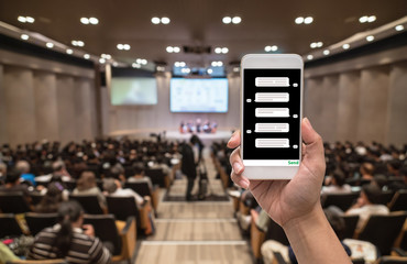 Female hand holding mobile phone showing chat message when Live via social network over Abstract blurred photo of conference hall or seminar room with attendee background, business technology concept