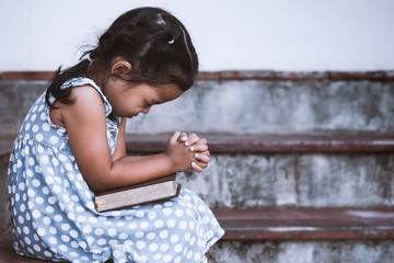 cute asian little girl closed her eyes and folded her hand in prayer on a holy bible for faith conce