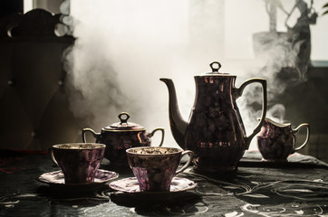 Food background tea and coffee theme. Old vintage ceramic tea or coffee pot with cups jug and sugar cup on dark background with lights and smoke. Silhouettes of ceramic tea pot and cup