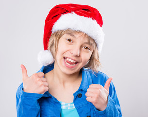 Wall Mural - Emotional portrait of little girl wearing Santa Claus red hat. Funny cute smiling child 10 year old thumbs up gesture and looking at camera on gray background. Winter holiday christmas concept.