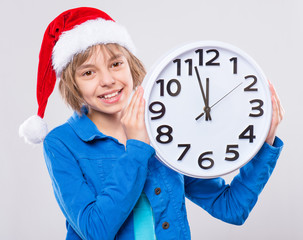 Wall Mural - Emotional portrait of attractive caucasian little girl wearing Santa Claus red hat. Funny cute smiling child 10 year old with big clock on gray background. Winter holiday christmas concept. 