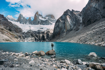 Wall Mural - One small man backpacker standing in front of a blue lake and mountains covered with snow