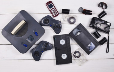 Vintage media and information technology. Entertainment 90s. Game console, gamepads, disks, audio cassettes, video cassettes, phone, film camera on a white wooden table. Top view. Flat lay. 
