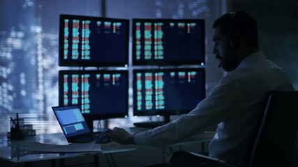 Leinwandbilder - Late at Night Trader Reads Numbers on His Multiple Displays with Stock Information on Them, He Also Consults Clients with Headset On. In Background Big City Window View. Shot on RED Cinema Camera 4K
