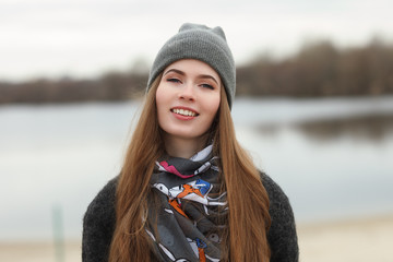 Full length lifestyle portrait of young and pretty adult woman with gorgeous long hair posing in city park with shallow depth of field in grey coat, hat, scarf