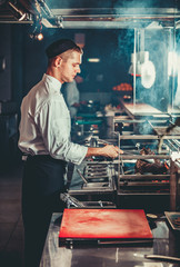 Wall Mural - Food concept. Young handsome chef in white uniform monitors the degree of roasting and turns meat with the forceps in interior of restaurant kitchen. Preparing traditional beef steak on barbecue oven.