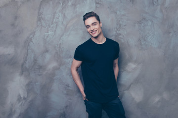 Attractive young guy is standing on the grey background. He is in black t shirt and jeans, smiling