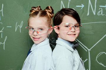 Boy and girl in white coats and protective eyeglasses standing together near chalkboard and smiling at camera, science school concept