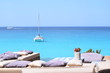 Luxuriöse blaue Yacht ankert im türkisfarbenen Wasser vor der Küste von Formentera, im Vordergrund Bänke und Kissen am Ufer, im Hintergrund ein Motorboot