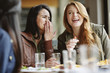 © Cavan Images - Happy woman whispering into friend's ear at table in restaurant
