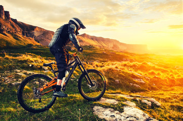 The rider in full protective equipment on the mtb bike is riding toward the sunset in the rays of the sunset sun against the background of the rocks of the setting sun and clouds