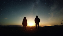 A Couple Of People Man And Woman Stand At The Sunset Of The Moon Under The Starry Sky With Bright Stars And A Milky Way. Silhouetted Photo Against The Starry Night Sky