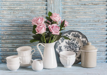 Pink Roses In A White Enameled Pitcher, Vintage Crockery On Blue Wooden Rustic Background. Kitchen Still Life In Vintage Style. Flat Lay