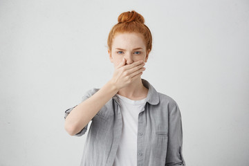 Indoor shot of serious young Caucasian woman with ginger hair wearing casual clothing covering mouth with hand as a sign of keeping secret or some confidential information, feeling frustrated