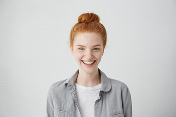 Wall Mural - Close up portrait of cute redhead European student girl with hair knot and clean freckled skin looking at camera with happy expression, having fun indoors, relaxing after morning lectures at college