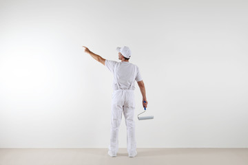 Wall Mural - Rear view of painter man pointing with finger the blank wall, with paint roller, isolated on white room